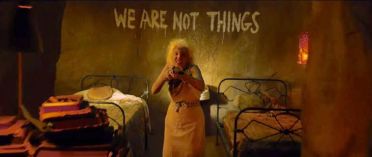 An old woman standing in an empty dorm points a shotgun at the viewer. On the wall behind her is scrawled the words “We are not things”