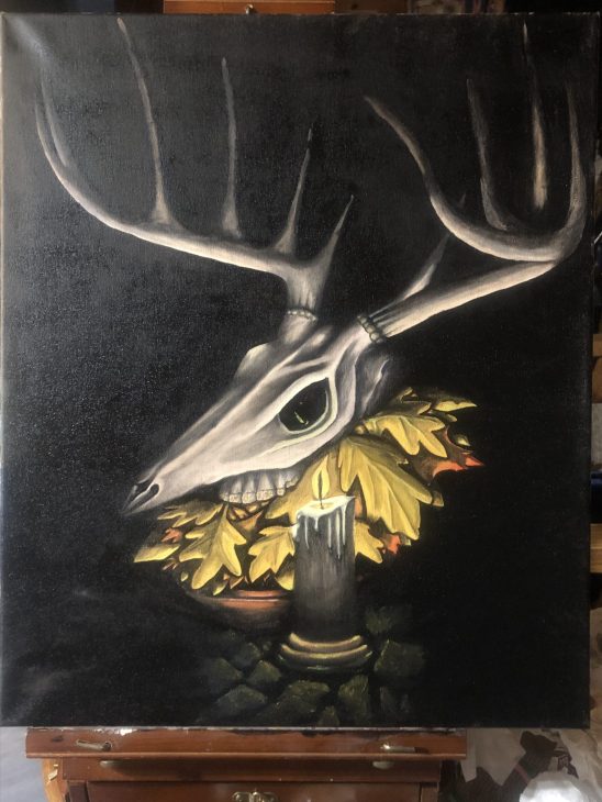 A stag skull sits on a bowl of autumnal leaves lit by a single candle against a black background. The candle is reflected in green light in the eye socket of the skull.