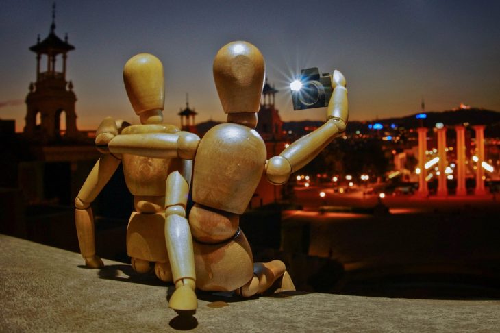 Two wooden anatomy dummies sit on a wall, one with an arm around the other’s shoulders, and take a selfie together.