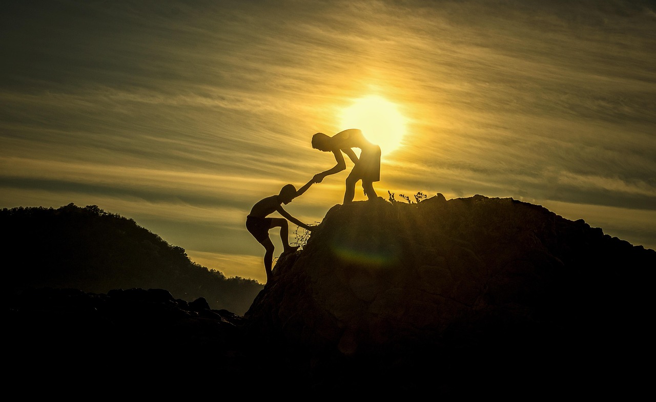 A young man helps another climb a short cliff face, silhouetted by a setting sun.